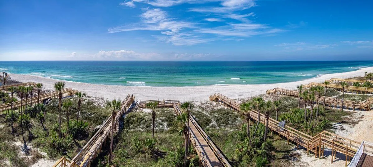 A serene, sun-dappled wooden walkway leading directly onto a private, palm-lined beach with gentle ocean waves in the distance. The view captures the exclusive beach access and the natural beauty surrounding the resort, inviting guests to stroll from their villa straight to the sand.