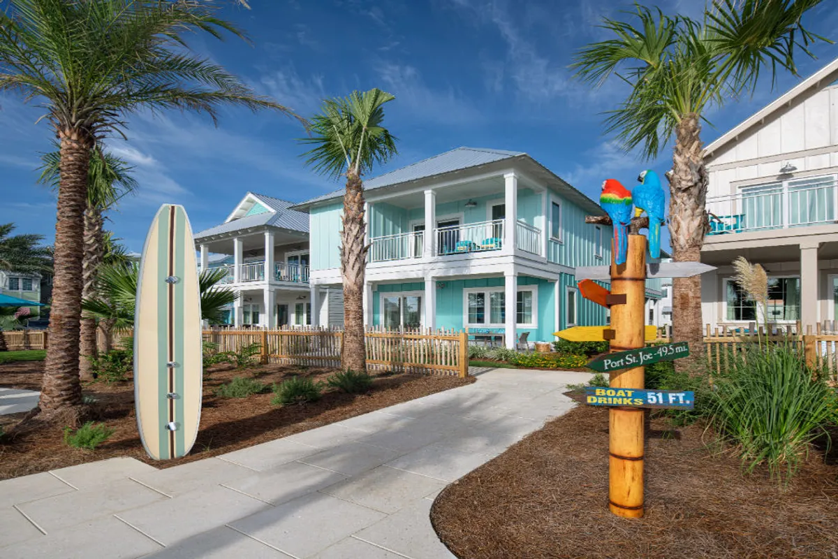A cheerful, laid-back exterior scene featuring a surfboard, a colorful signpost with directions, tropical palm trees, colorful parrots, and a glimpse of the house's architecture, including a balcony. The setting hints at opportunities for morning coffee near the surfboard, sunset views from the balcony, and evening gatherings.