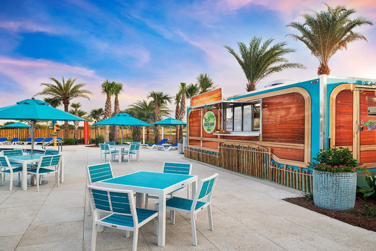 An inviting outdoor setting featuring a food truck surrounded by outdoor seating areas shaded by umbrellas and framed by palm trees. Guests can be seen enjoying meals and socializing in a lively, poolside atmosphere that suggests relaxation and leisure under the Gulf sun.