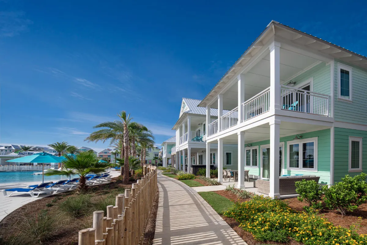 An exterior shot capturing a lively pool area surrounded by greenery, colorful building facades, and porch or patio spaces. Guests are seen socializing, swimming, and relaxing on porches, suggesting options for enjoying morning coffee or unwinding at sunset in an outdoor setting.