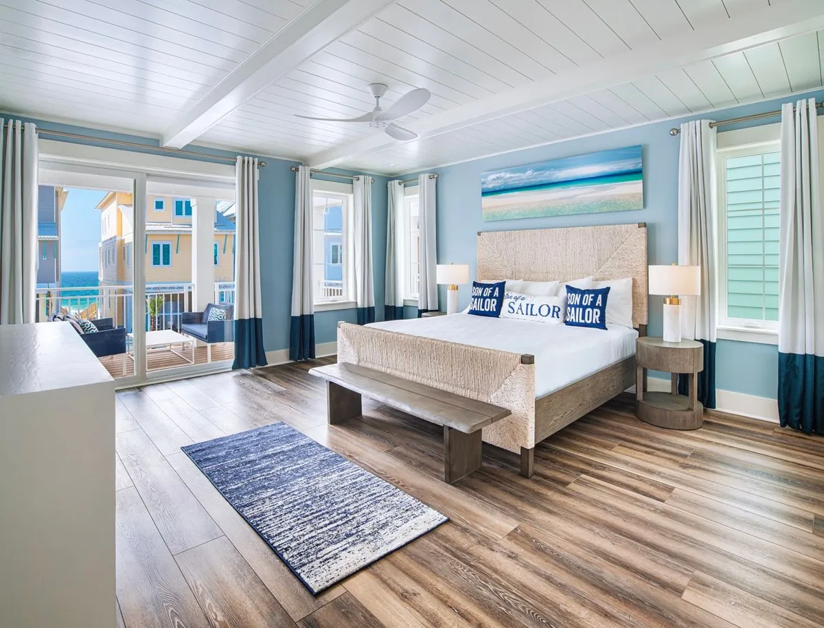 A serene bedroom featuring a cozy bed with sailor-themed pillows, large windows offering a direct ocean view, decorative sea-inspired artwork, and warm wooden flooring. The room is filled with natural light, creating a tranquil, inviting atmosphere perfect for relaxation and unwinding after a day of coastal activities.