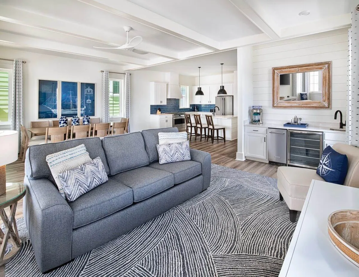 A bright, open-plan living space featuring a plush sofa with decorative pillows, a spacious dining table with chairs, a large kitchen island with stools, and a bar area equipped with modern appliances. The area is accented with framed artwork and bathed in natural light, creating a warm, inviting atmosphere ideal for socializing and gathering.
