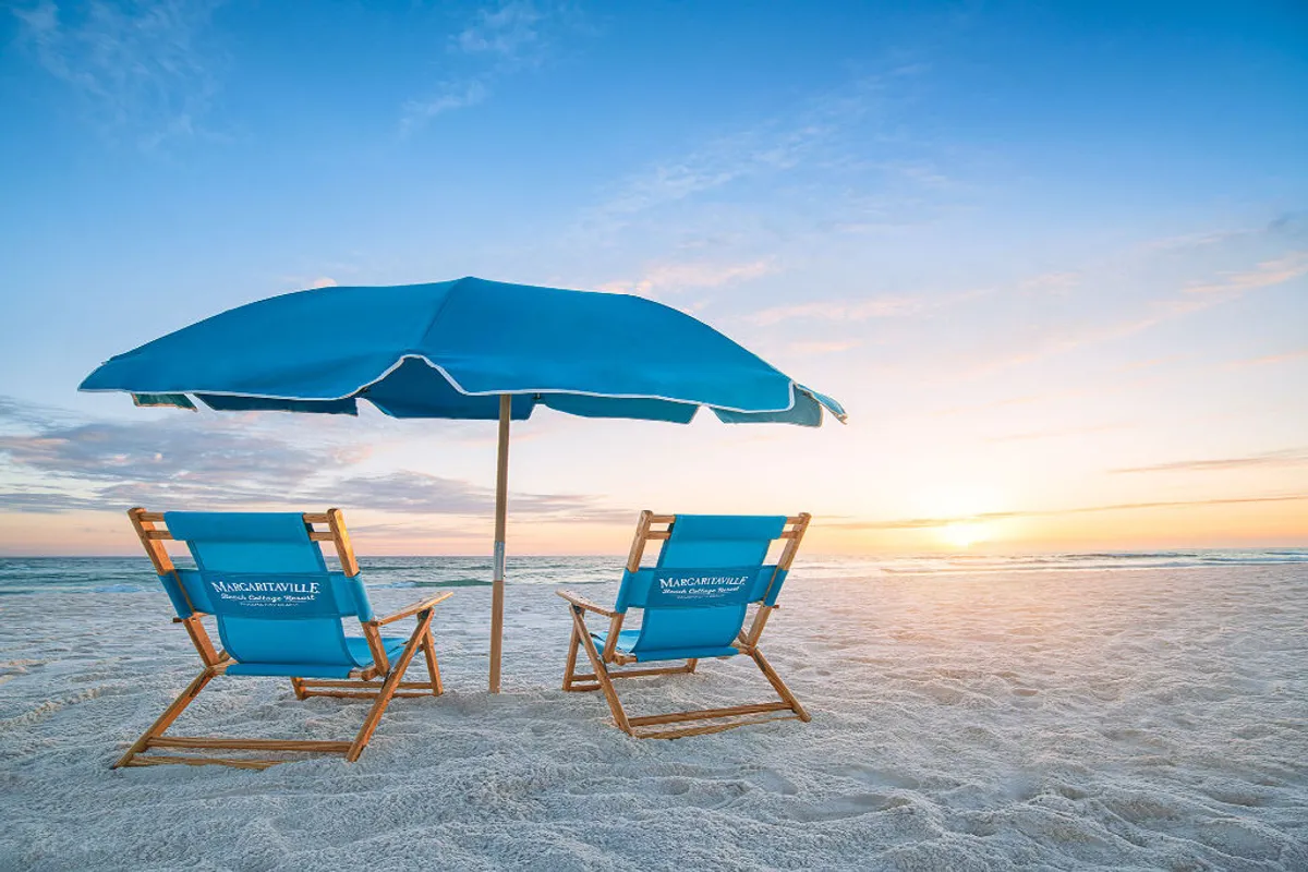 The image showcases two inviting blue wooden beach chairs with Margaritaville branding under a large blue umbrella, set on a pristine, expansive sandy beach as the sun sets over the shimmering Gulf of Mexico. The tranquil scene evokes relaxation and exclusivity, with the ocean horizon glowing in golden hues.