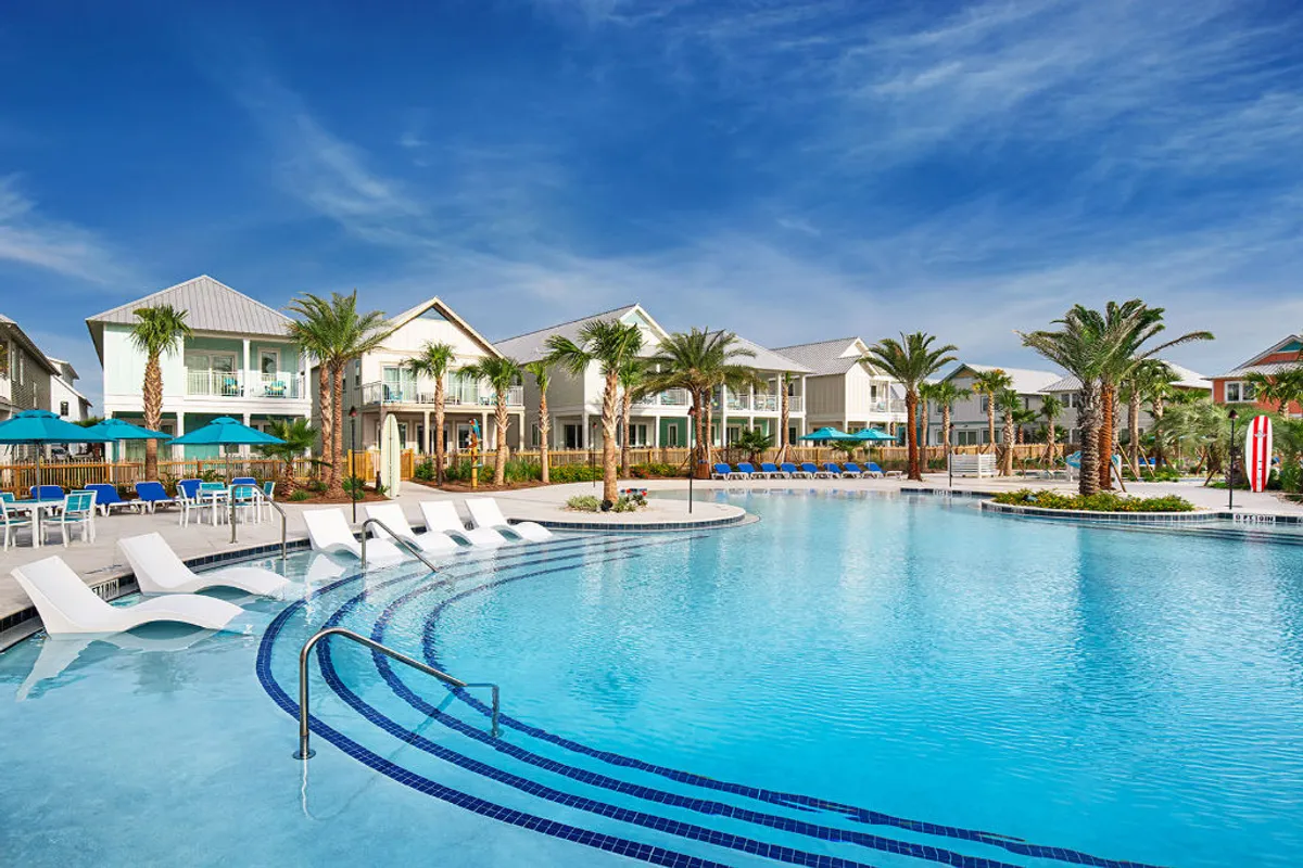 A sprawling resort-style swimming pool surrounded by palm trees, with modern coastal homes visible in the background. The pool is dotted with lounge chairs and umbrellas, creating inviting spaces for groups to gather, swim, and relax. The landscaped gardens and open layout exude a vacation-ready ambiance, while hints of the Gulf can be glimpsed from the balconies.