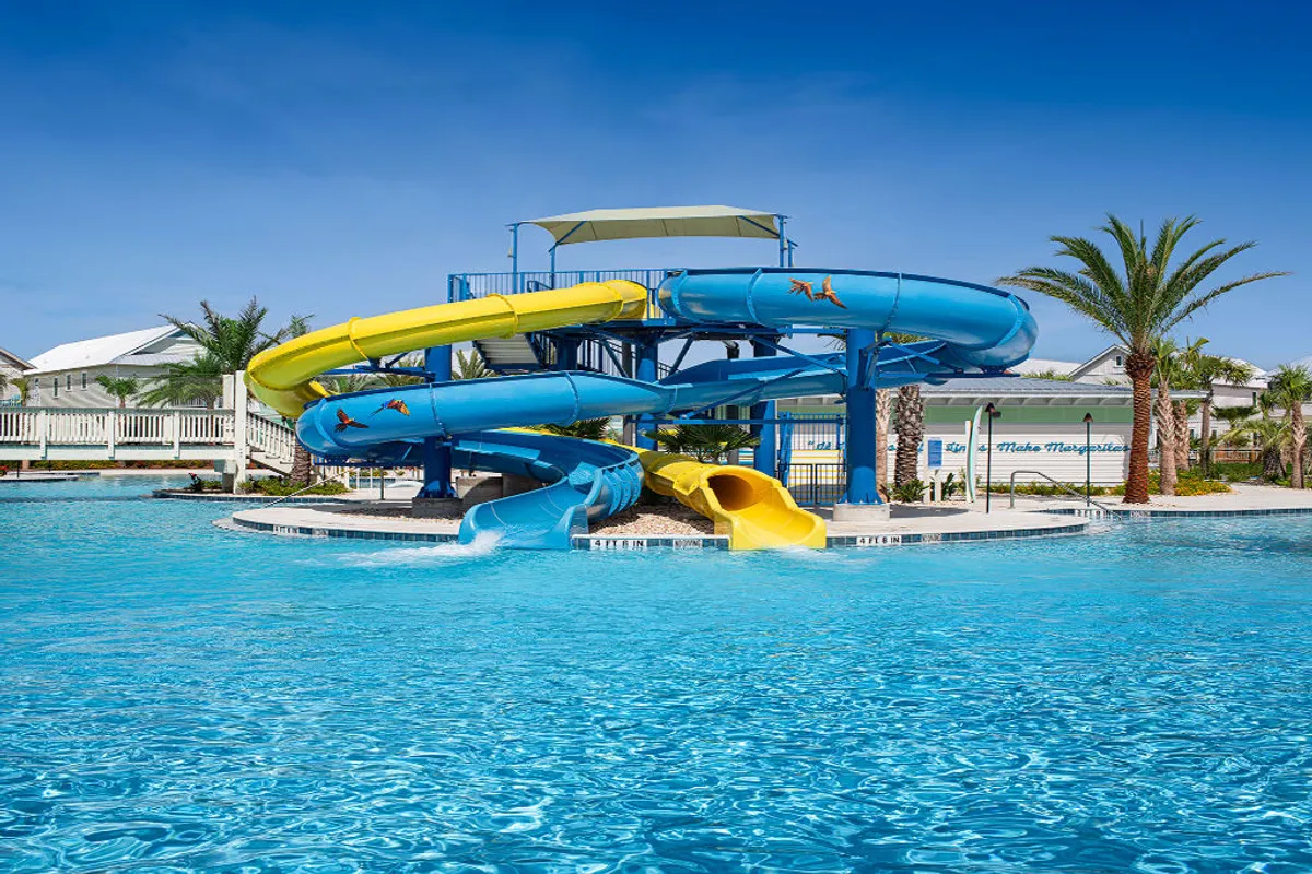 A vibrant scene of the resort’s large heated pool surrounded by palm trees, featuring multi-colored blue and yellow waterslides. The pool area is bordered by white fencing and resort buildings, and the lively atmosphere is enhanced by visible families and children enjoying watersliding, swimming, and relaxing poolside.