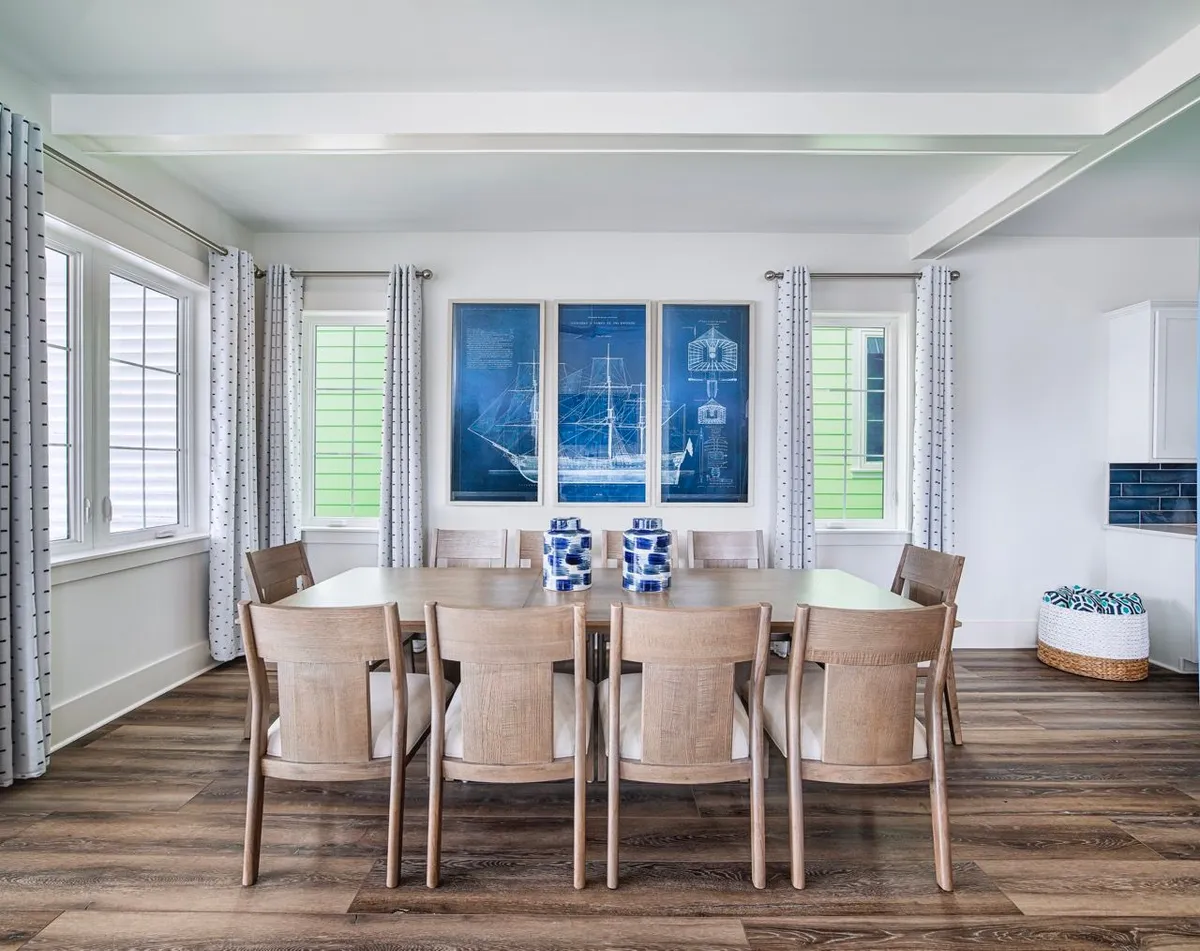 This interior image features a large wooden dining table that seats eight, set in a bright, airy room with nautical-themed blue artwork and decorative accents. Sunlight streams through patterned curtains, creating a welcoming space that radiates comfort and coastal charm.