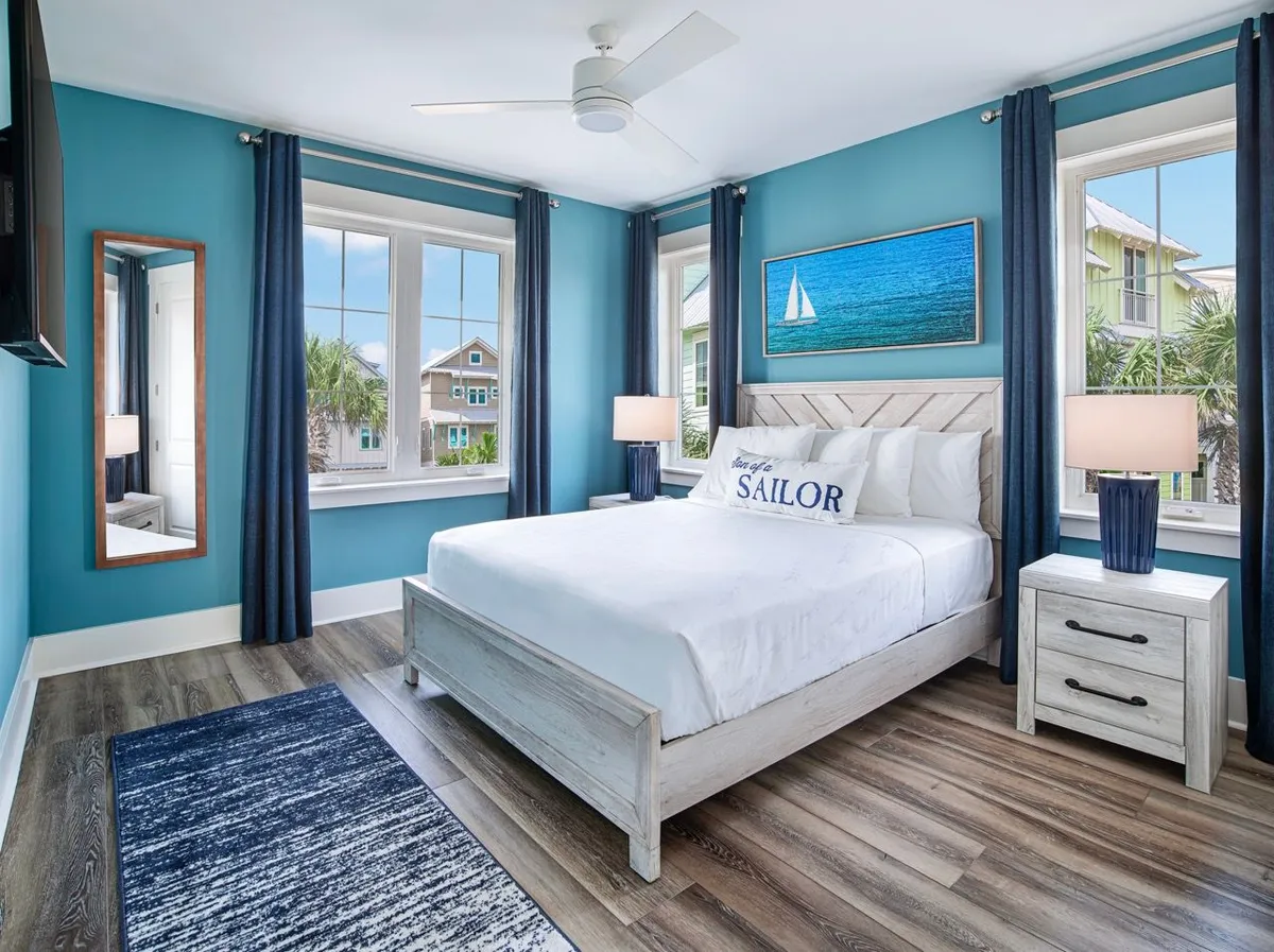 A calm, nautically-themed bedroom featuring a king-sized bed with a playful 'Sailor' pillow, matching blue lamps on nightstands, nautical sailboat artwork, and expansive windows that provide views of neighboring coastal homes and swaying palm trees.